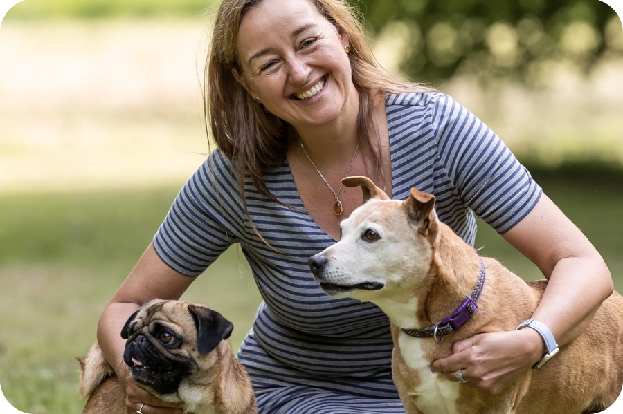 Smiling woman kneeling on grass with arms around dogs
